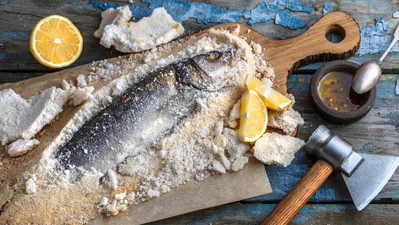 Fisch liegt in einer Salzkruste und Zitronen auf einem Holzbrett, daneben ein Klopfer. © PantherMedia Foto: kopachinsky