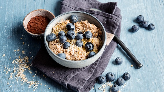 Eine Schüssel mit Frühstücksbrei und Blaubeeren und ein Schälchen mit Rohkakao stehen auf einem Tisch. © NDR Foto: Claudia Timmann