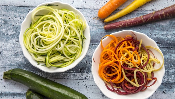 Eine Schüssel mit Zucchninistreifen steht neben einer mit Karottenstreifen, daneben liegen eine ganze Zucchini und verschieden farbige Karotten. © imago/Westend61 Foto: Sandra Roesch