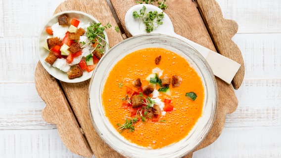 Ein Suppenteller mit gekühlter Gemüsesuppe mit Feta steht auf einem Brett. Daneben steht ein Tellerchen mit Croutons und Feta. © NDR Foto: Claudia Timmann