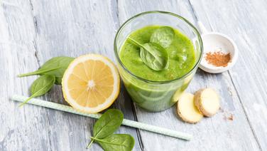 Ein Glas mit grünem Smoothie steht auf einem Tisch. Daneben liegt verstreut Spinatblätter, eine Zitronenhälfte, Ingwerscheiben und ein Schälchen Zimt. © NDR Foto: Claudia Timmann
