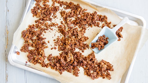 Auf einem Holztisch liegt ein mit Backpapier ausgelegtes Blech. Darauf befindet sich ein Knusper-Müsli aus Banane, Getreide, Körnern und Mandeln. © NDR Foto: Claudia Timmann