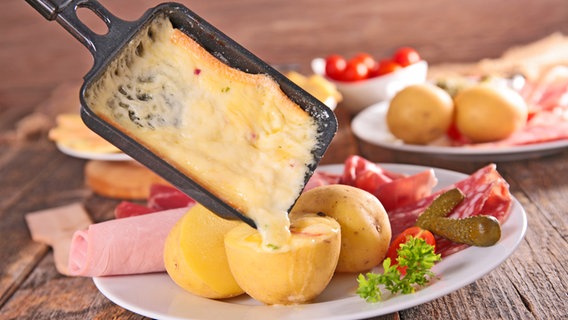 Geschmolzener Raclette-Käse fließt aus einem Pfännchen auf einen Teller mit Kartoffeln und Wurst © M.studio - Fotolia 