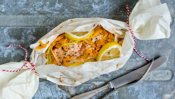 Auf einem Tisch liegen ein Päckchen gefüllt mit Lachs und Gemüse sowie ein Besteck. © NDR Foto: Claudia Timmann