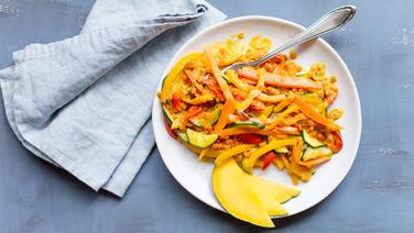 Auf einem Tisch steht ein Teller Gemüsecurry mit roten Linsen und in Spalten geschnittener Mango. Daneben liegt eine Stoffserviette. © NDR Foto: Claudia Timmann
