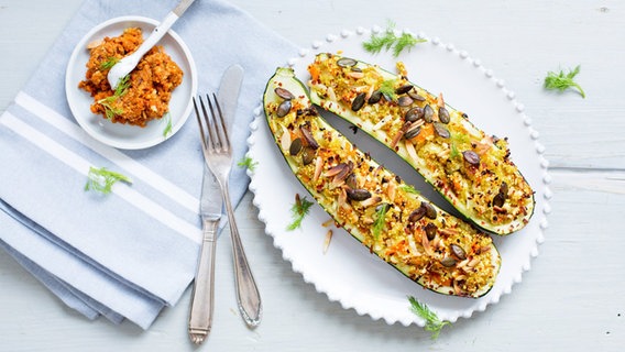 Auf einem Tischtuch mit Serviette und Besteck stehen Ofen-Zucchini mit Gemüse-Quinoa und Kernen. In einem Extra-Schälchen ist noch weitere Gemüsefüllung. © NDR Foto: Claudia Timmann