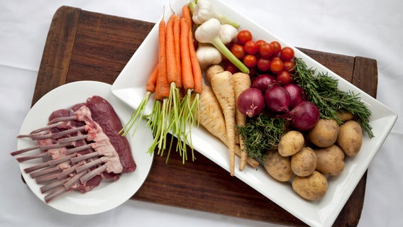 Lammkarree, Karotten, Knoblauch, Pastinaken, Rosmarin, Zwiebeln, Tomaten und Kartoffeln auf einem Holzbrett. © NDR Foto: Claudia Timmann