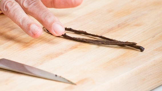 Eine Hand greift nach zwei Vanille-Schoten auf einem Holzbrett. © NDR Foto: Claudia Timmann