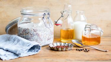 Ein Weckglas mit Overnight Oats, eine Flasche Milch, ein Glas Honig, eine Flasche Leinöl, ein Löffel Kurkuma und ein Schälchen Kakaopulver auf einem Brett. © NDR Foto: Claudia Timmann