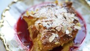 Brotpudding mit Puderzucker, Mandelsplittern und Himbeersoße auf einem Teller serviert. © NDR 