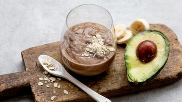 Ein Frühstücksshake auf einem Holzbrett, daneben eine halbe Avocado. © NDR Foto: Claudia Timmann