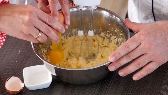 Hände schlagen ein Ei in eine Teigmischung in einer Rührschüssel. © NDR Foto: Claudia Timmann