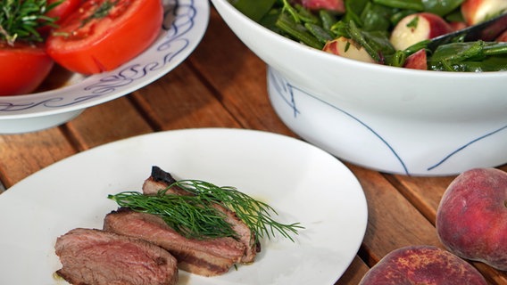 Gegrillte Lammhüfte, Bohnen-Pfirsich-Salat und gegrillte Tomaten auf einem Teller und in Schüsseln angerichtet. © NDR Foto: Florian Kruck