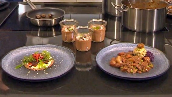 Auf einer Arbeitsplatte stehen Pilzragout mit Knödeln, Blini mit Paprika und Salat und drei Gläser mit Mousse au Chocolat. © NDR 