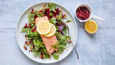 Salmon fillet lies on a plate with a salad, next to it a bowl of pomegranate seeds and half an orange.  © NDR Photo: Claudia Timmann