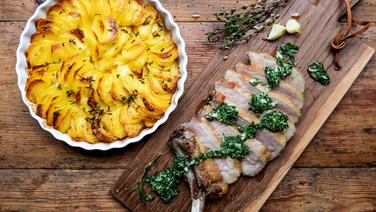 Glasierte Schweinekoteletts auf einem Brett serviert und Kartoffelgratin in einer Auflaufform. © NDR Foto: Claudia Timmann