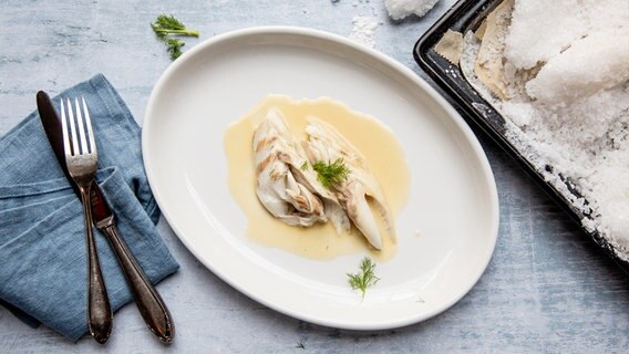 Ein Stück Wolfsbarschfilet mit Soße auf einem Teller, daneben ein Backblech mit Salzkruste. © NDR Foto: Claudia Timmann