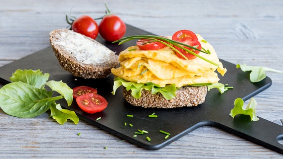 Ein Holzbrett mit Roggenbrötchen, Rührei und Tomaten steht auf dem Tisch. © NDR Foto: Claudia Timmann