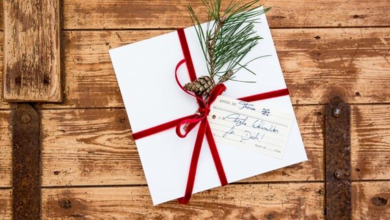 Ein quadratischer weißer Geschenkkarton mit rotem Samtband, einem Kiefernzweig und einem Zapfen. © NDR Foto: Claudia Timmann