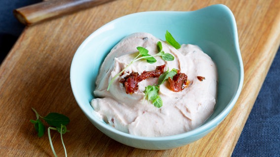 A bowl of tomato dip and a small knife lie on a wooden board.  © NDR Photo: Claudia Timmann