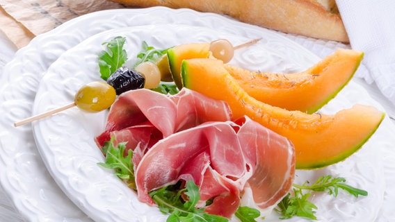 Auf einem Teller liegen dünne Scheiben Parmaschinken sowie Oliven und Melonenspalten. © imago images Foto: Panthermedia