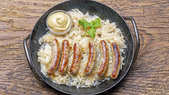 Bratwürstchen und ein Schälchen Senf in einer Pfanne mit Sauerkraut. © imago images / Panthermedia 
