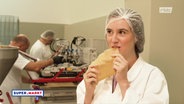 Frau in Lebensmittelfabrik mit Hygienehaube beißt in eine dünne cremefarbene Platte, im Hintergund arbeiten zwei Männer an Maschinen, oben rechts im Bild das Logo des rbb, unten links der Sendungstitel Super.Markt © rbb 