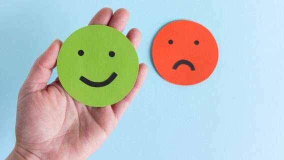 Ein trauriger und ein fröhlicher Smileys. Der fröhliche Grüne liegt in einer Hand, der traurige Rote liegt auf einem hellblauen Hintergrund © photocase Foto: Marie Maerz