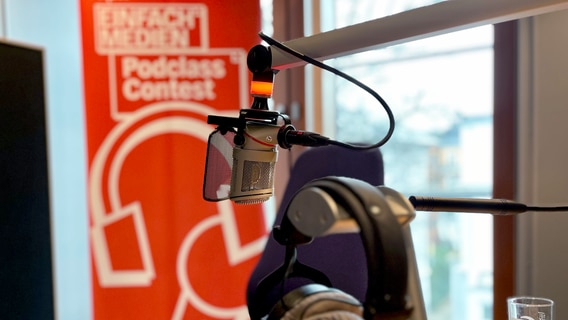 Ein Mikrofon und Kopfhörer. Dahinter ein Banner vom NDR einfach.Medien Podclass Contest. © NDR 