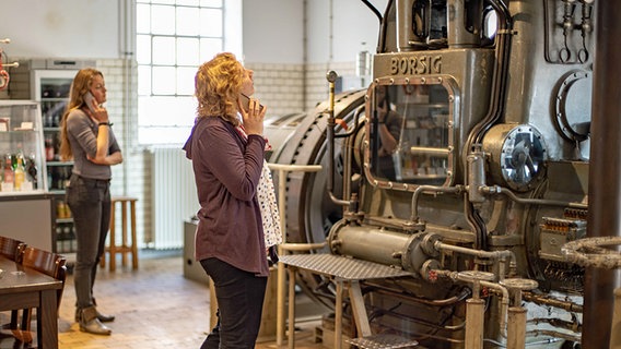 Dua pengunjung dengan pemandu audio berpose di depan mesin uap di Museum Industri Elmshorn.  © Museum Industri Elmshorn Foto: Peter Eberle