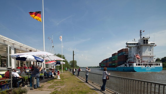 Menschen stehen an der Schiffsbegrüßungsanlage in Rendsburg und schauen auf den Nord-Ostsee-Kanal, auf dem ein Containerschiff vorbeifährt. © imago images/Hoch Zwei Stock/Angerer 