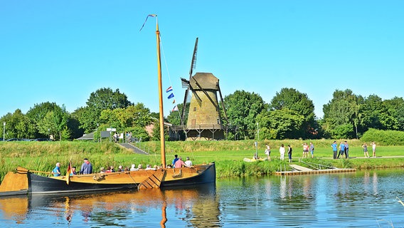 Eine Vechtezompe liegt am Ufer der Vechte, im Hintergrund eine historische Mühle und Urlauber. © Samtgemeinde Emlichheim, 