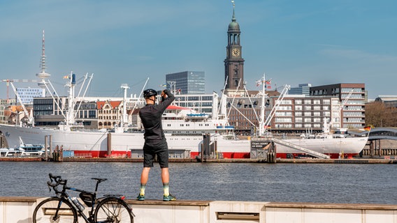 Ein Radfahrer steht am Ufer der Elbe und fotografiert das Hamburger Hafenpanorama mit dem Michel und dem Museumsschiff "Cap San Diego". © picture alliance /dpa Foto: Markus Scholz