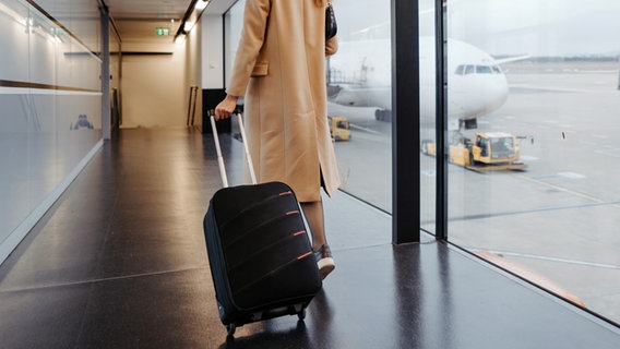 Eine Frau geht mit einem kleinen Rollkoffer einen Gang in einem Flughafen entlang. © Colourbox Foto: Big Shot Theory