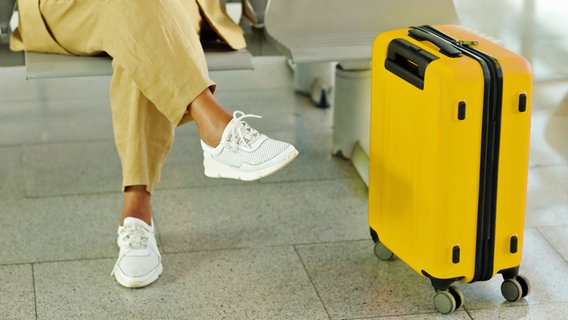 Eine Frau sitzt mit einem gelben Rollkoffer auf einer Bank in einem Flughafen. © Colourbox Foto: Andrii