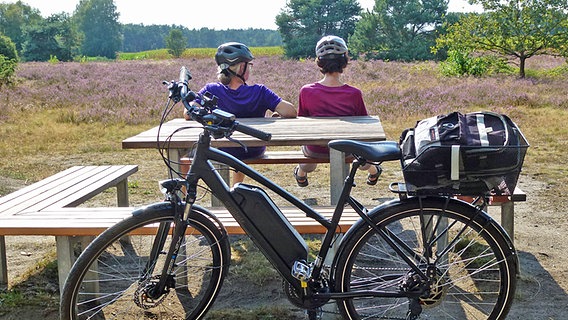 Zwei Frauen mit Fahrradhelmen sitzen auf einer Bank und blicken auf eine Heidelandschaft. © NDR Foto: Axel Franz