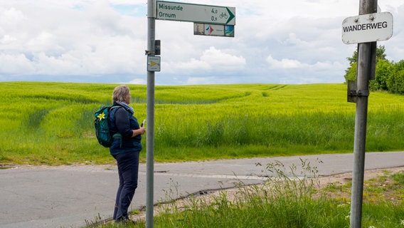 Eine Frau steht an einer Abzweigung an einer Straße, mehrere Hinweisschilder sind an Pfosten befestigt. Im Hintergrund sind Getreidefelder zu erkennen. © NDR Foto: Anja Deuble