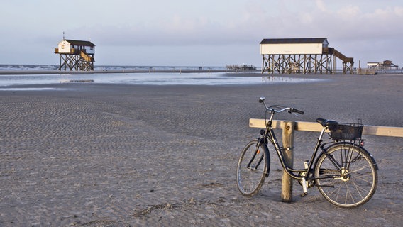 Ein Fahrrad lehnt einem Holzständer am Strand von St. Peter-Ording, im Hintergrund sind Stelzenhäuser zu sehen. © picture alliance/chromorange Foto: Monika Wirth
