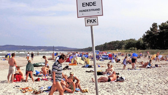 Besucher an einem FKK-Strand bei Binz auf Rügen sowie ein Hinweisschild auf den FKK-Strandabschnitt. © imago images Foto: Jens Koehler