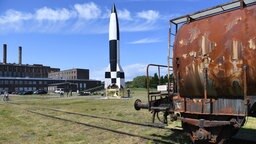 Ein verrosteter Eisenbahnwaggon steht im Freigelände des Historisch-Technischen Museums Peenemünde, dahinter ist der Nachbau einer V2-Rakete zu sehen.