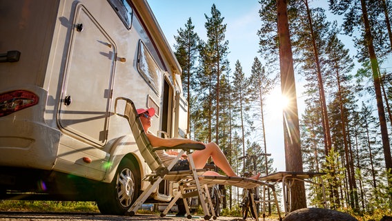 Eine Frau sitzt auf einem Liegestuhl vor einem Wohnmobil im Wald. © Colourbox Foto: Andrey Armyagov