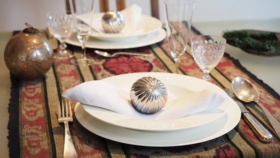 Auf einem orientalischen Tischläufer stehen weiße Teller mit weißen Stoffservietten und silbernen Kugeln. © NDR Foto: Anja Deuble