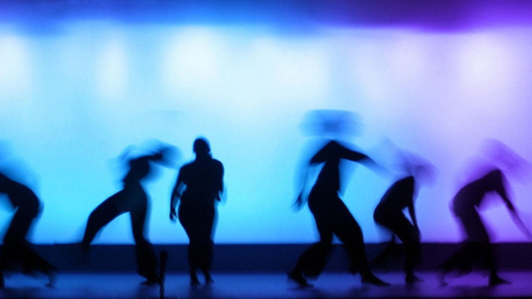 Tanzende Figuren vor blau-lila beleuchtetem Hintergrund.