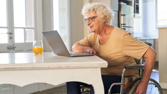 Eine ältere Frau sitzt lächelnd in einem Rollstuhl an einem Küchentisch und schaut auf einen Laptop. © picture alliance / Zoonar Foto: Robert Kneschke