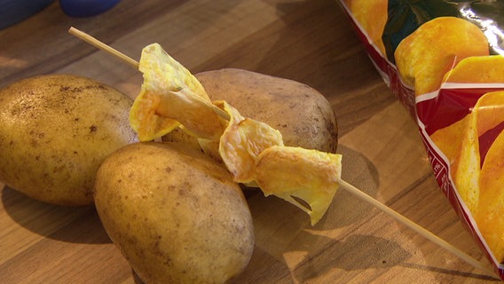 Selbstgemachte Chips aus frischen Kartoffeln auf einem Holzstäbchen, daneben Chipstüte aus dem Supermarkt. © NDR 