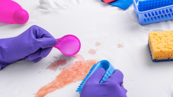 Hände mit Gummihandschuhen, Bürste und Dosierlöffel über einem weißen T-Shirt mit Fleck © colourbox 