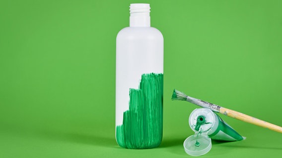 Eine weiße Flasche steht vor einem grünen Hintergrund. Die Flasche wurde teilweise mit grüner Farbe angestrichen. Pinsel und Farbtube liegen daneben. © picture alliance / Zoonar Foto: Firn