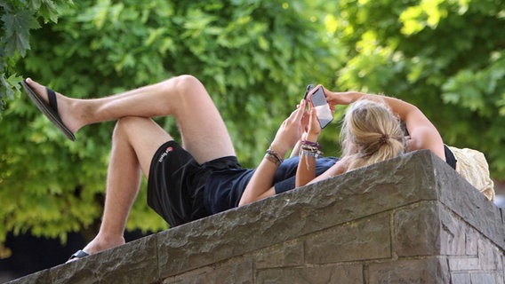 A couple lies on a wall in the shade under trees, both holding smartphones.  © imago images Photo: Ralph Peters