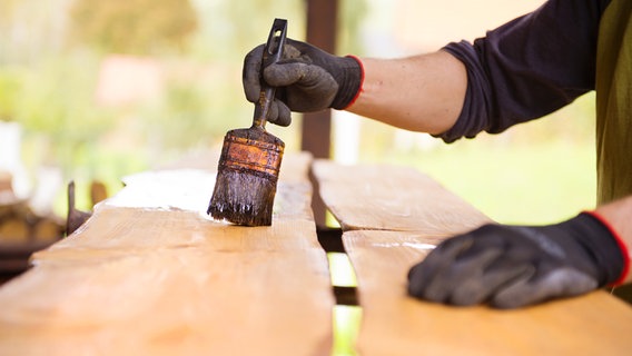 Eine Person mit Handschuhen lackiert ein längliches Stück Holz. © Colourbox 