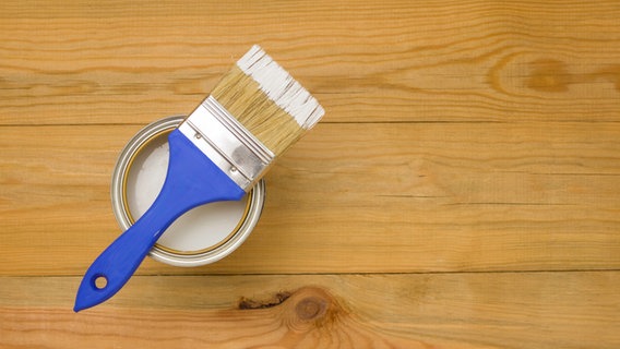 Ein Pinsel liegt quer auf einer Dose mit weißem Holzlack, darunter eine hölzerne Fläche. © Colourbox Foto: -